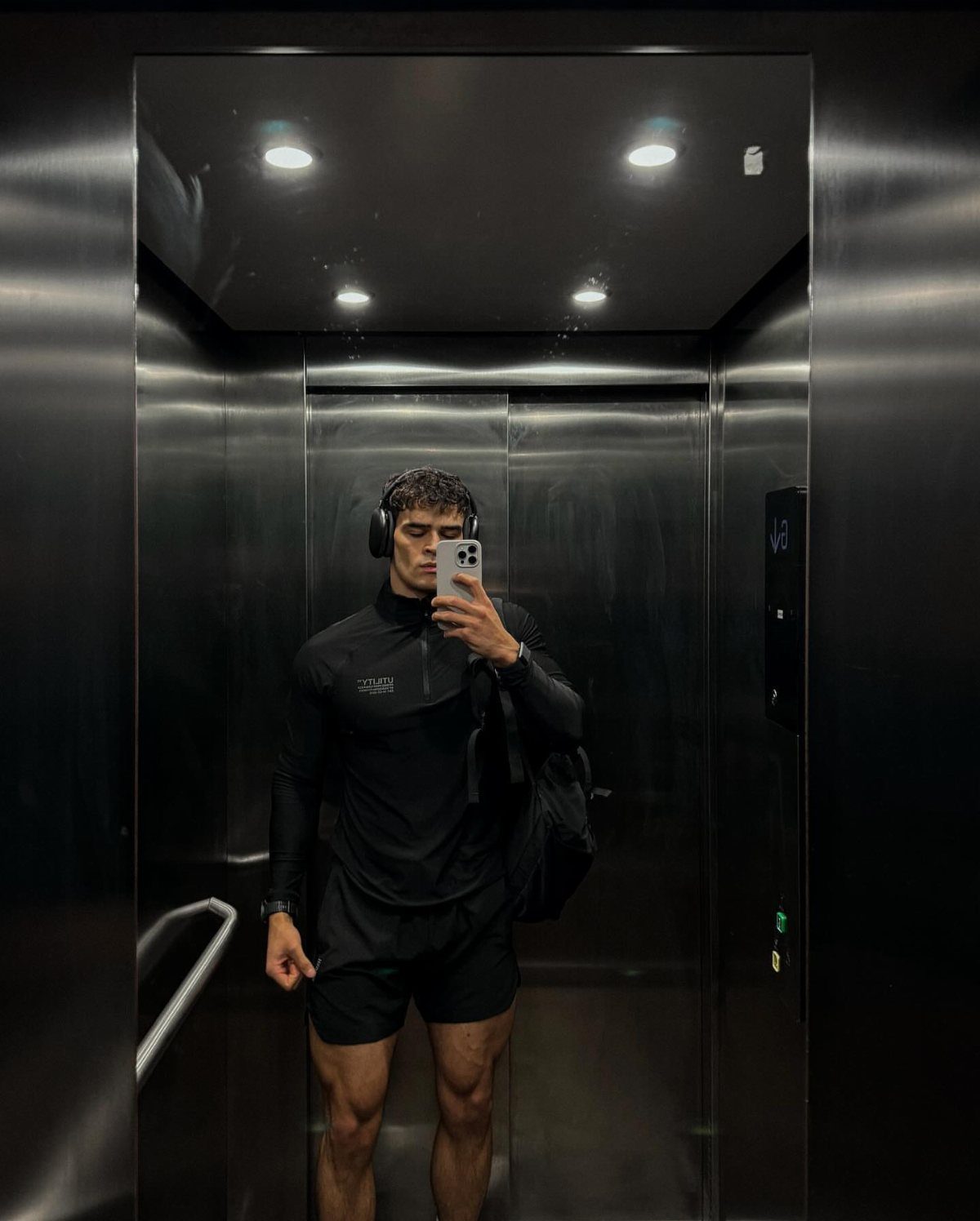 Man in black athletic wear taking a mirror selfie inside a metallic elevator.
