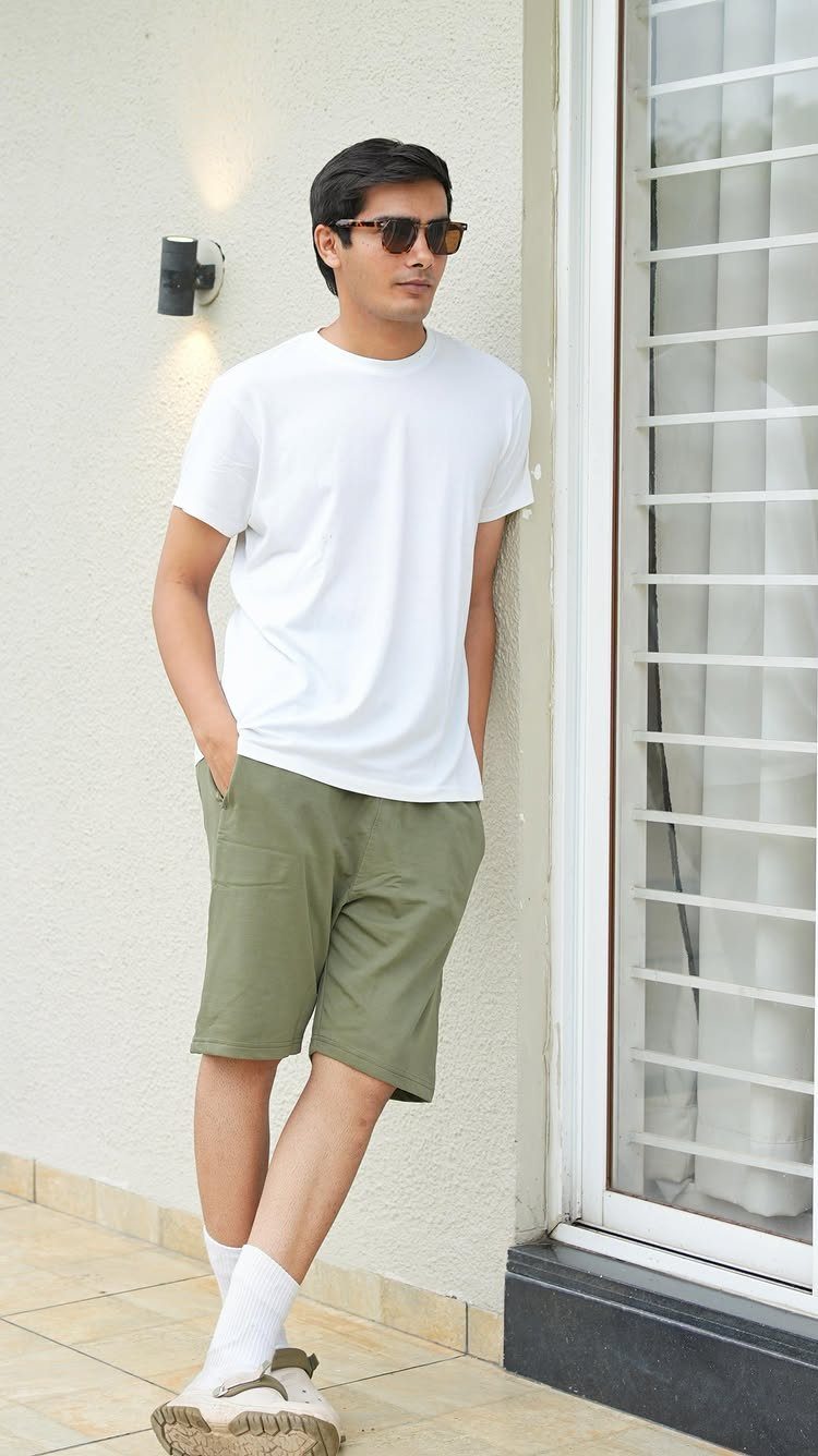 Man in white t-shirt and olive green shorts leaning against a white wall