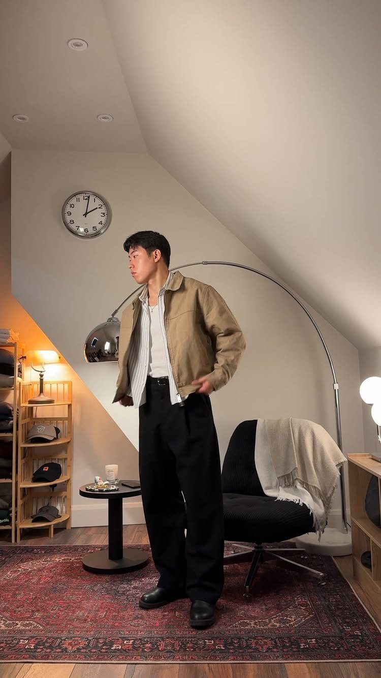 Man wearing a tan jacket and loose black trousers standing in a room