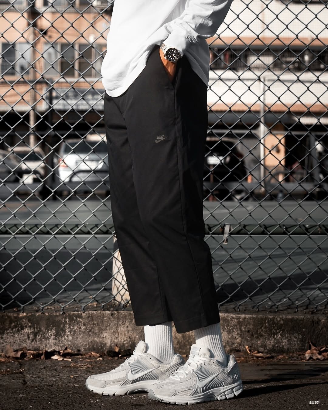 Person wearing black cropped trousers, white socks, and white sneakers by a fence.