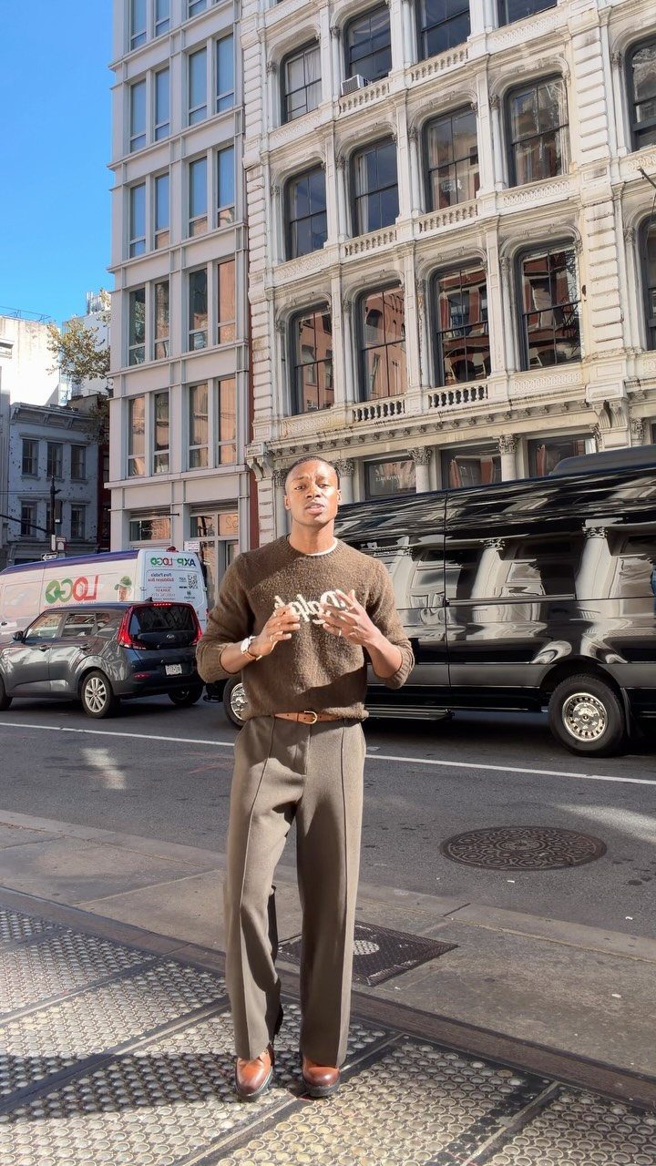 Man wearing olive green trousers and brown sweater standing on a city street.
