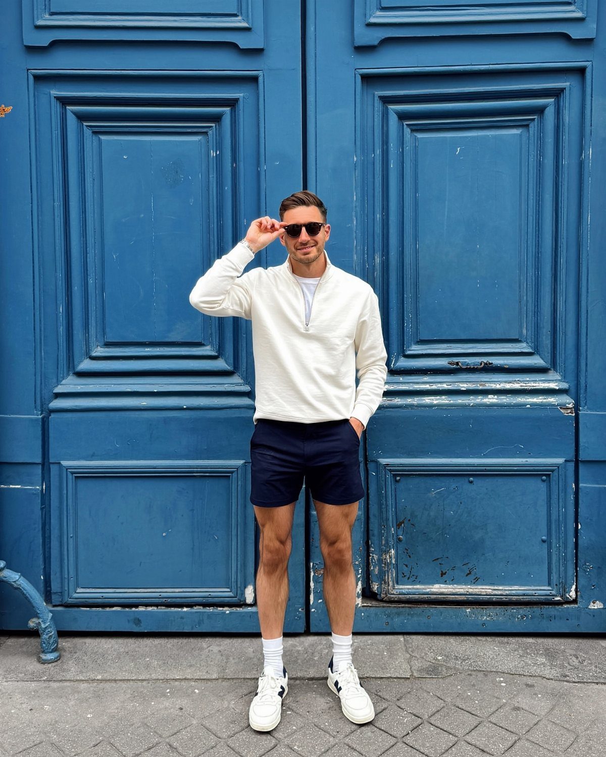 Man wearing white pullover and dark blue shorts standing before blue doors.
