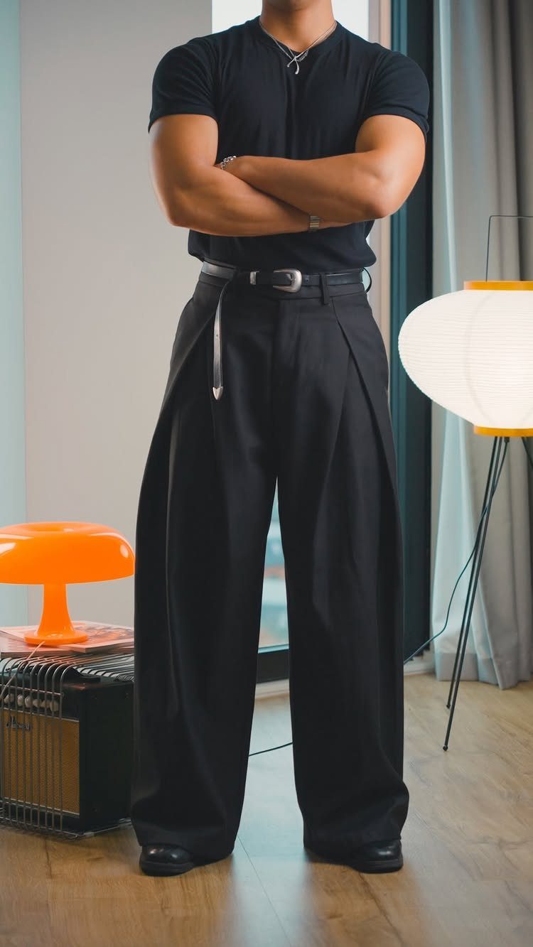 Man wearing a black short-sleeved shirt and wide-leg black pleated trousers standing indoors.