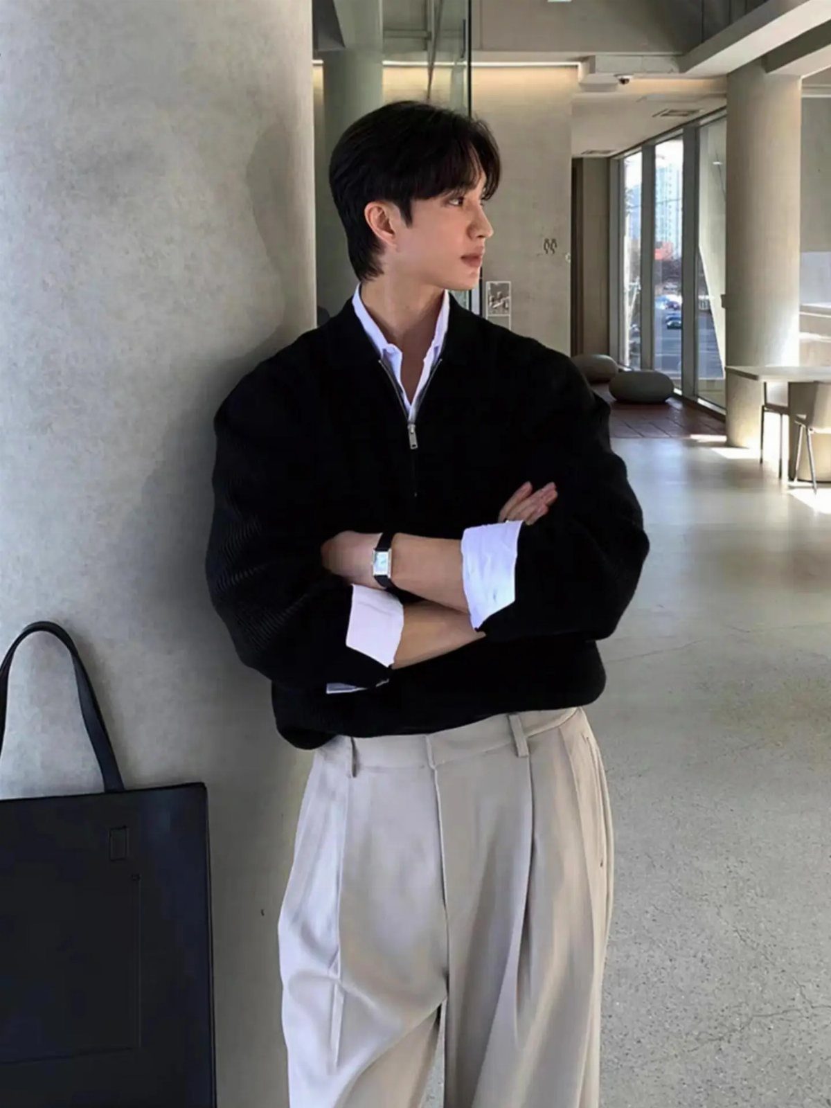 Man wearing a black sweater over a white shirt and beige trousers.