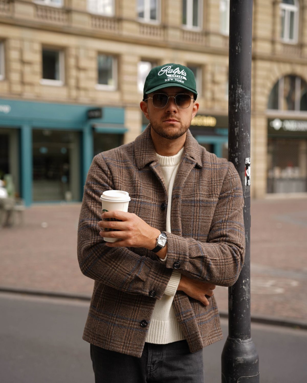 Man in brown plaid overshirt and white sweater holding coffee on city street