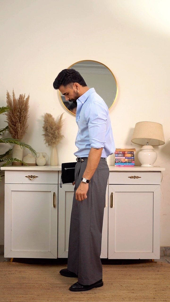 Man wearing wide-leg grey trousers and a light blue shirt standing indoors.