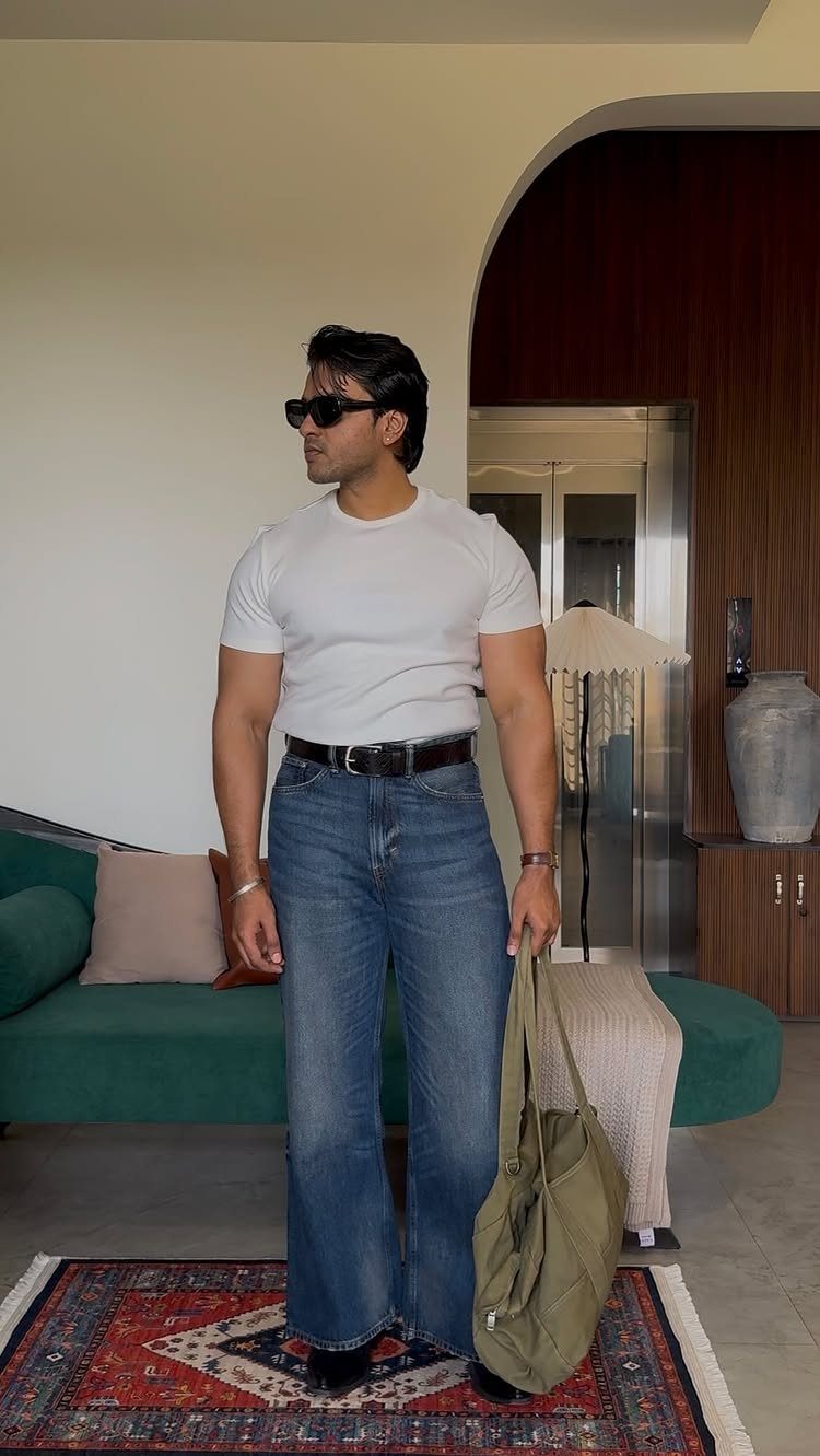 Man wearing a white t-shirt and wide-leg blue jeans holding a green tote bag.