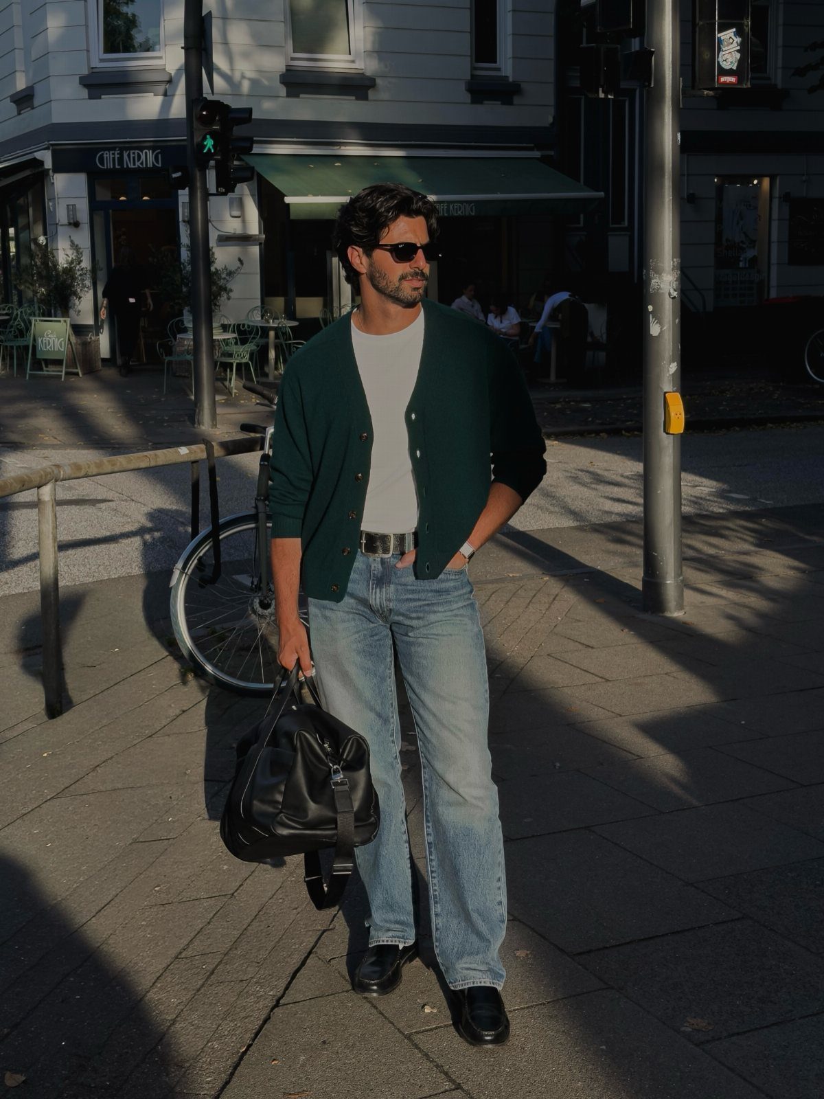Man in dark green cardigan and light blue jeans standing on city street