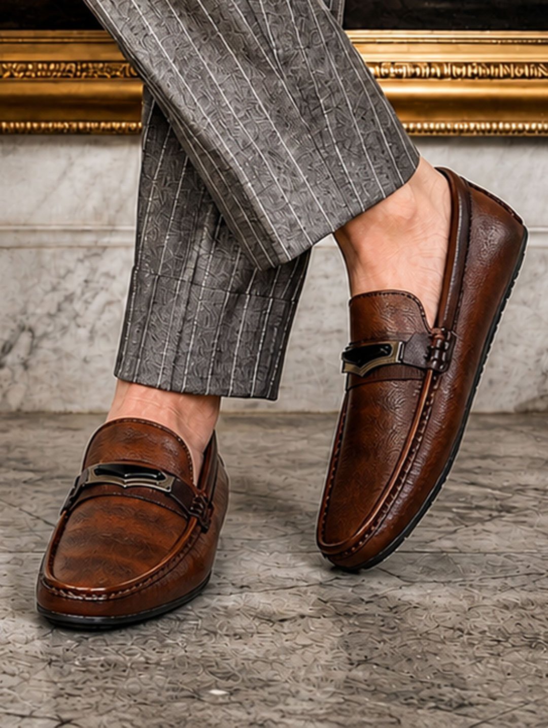 Brown leather loafers with metal accents paired with grey pinstriped trousers.