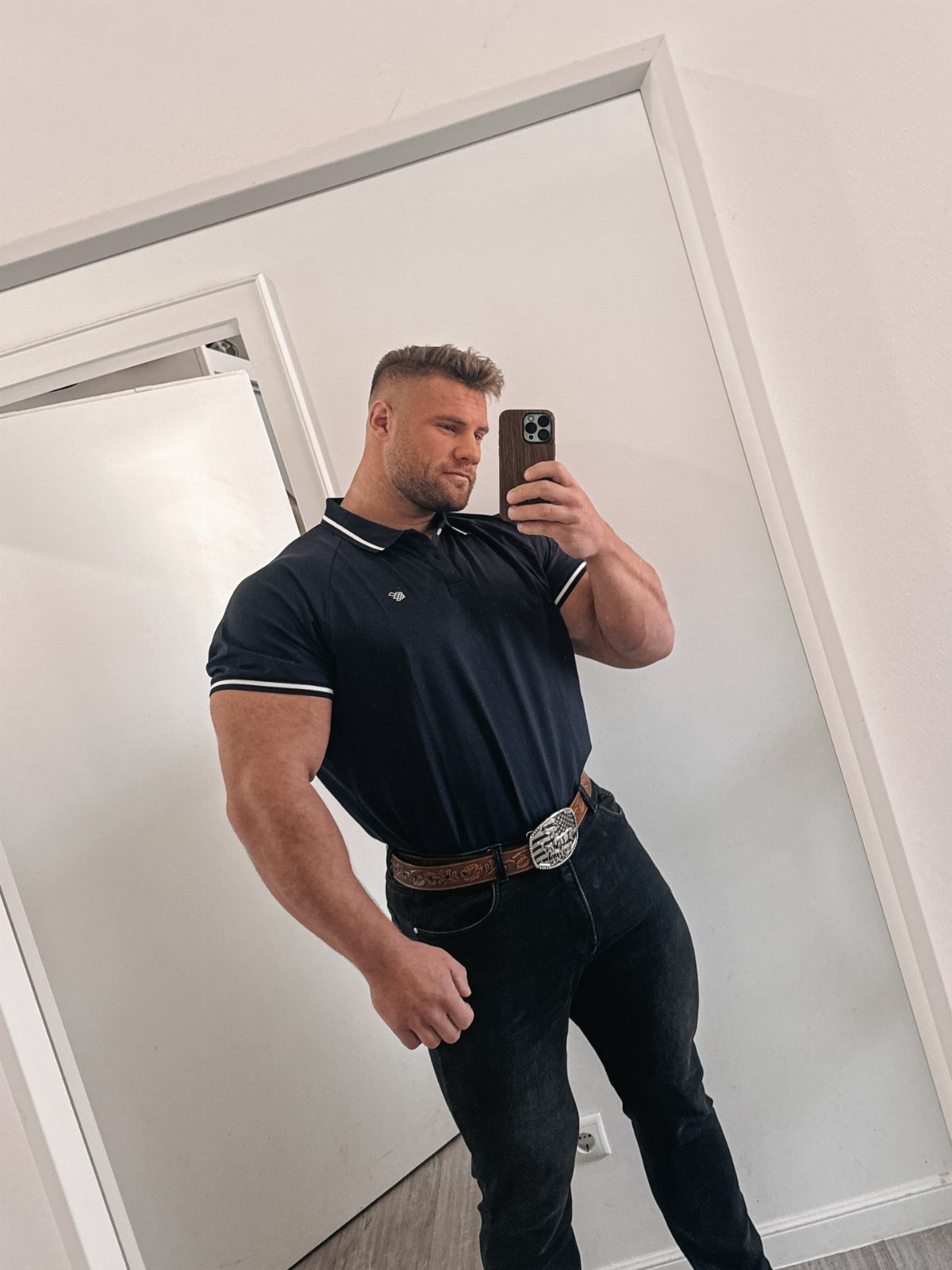 Muscular man in navy polo shirt and black jeans taking a mirror selfie.