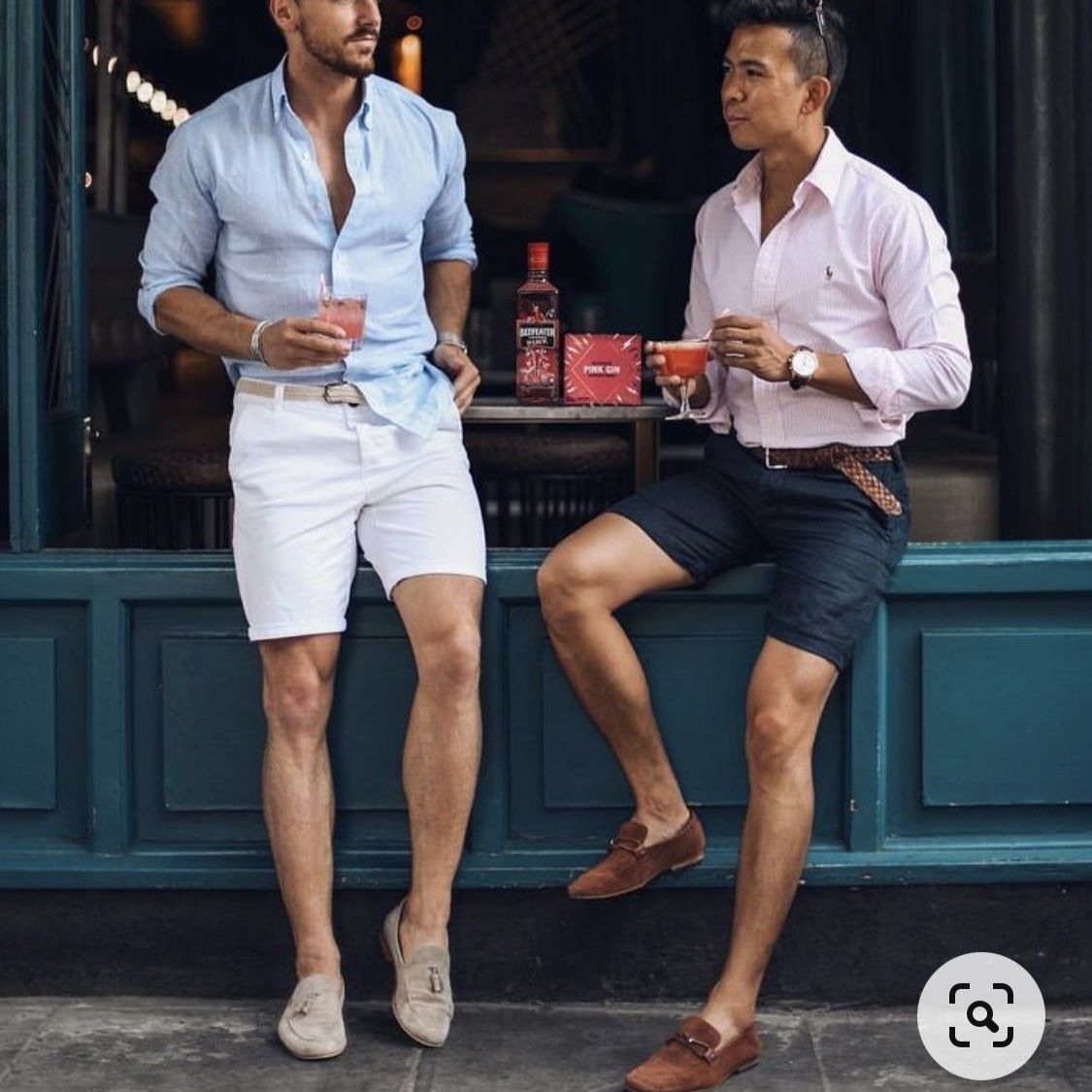 Two men in casual shirts and shorts holding drinks outdoors by a wall