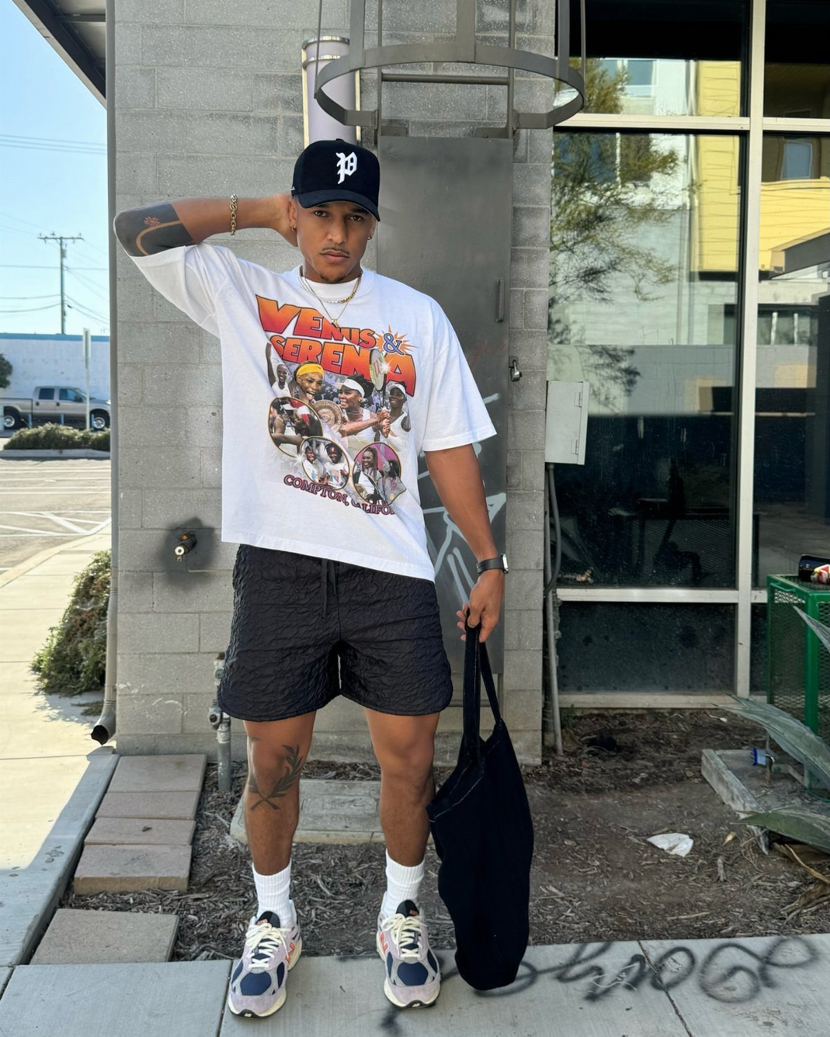 Man wearing a white graphic tee, black shorts, and chunky sneakers standing outdoors.
