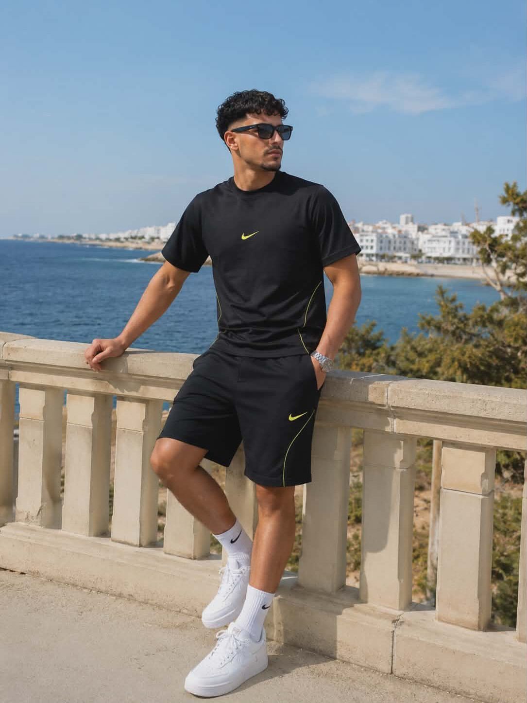 Man in black athletic short set and white sneakers leaning on stone railing.