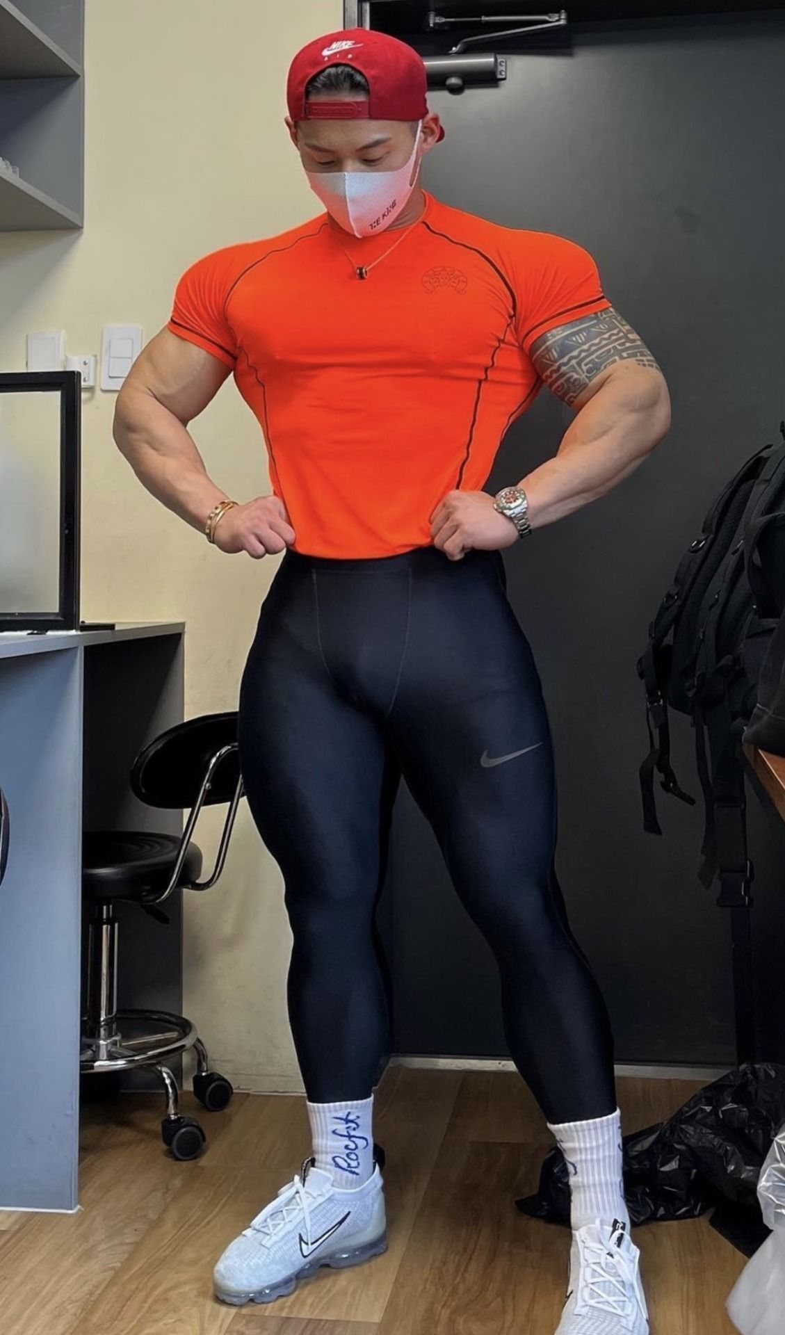 Muscular man in orange athletic shirt and black tight workout leggings posing indoors.