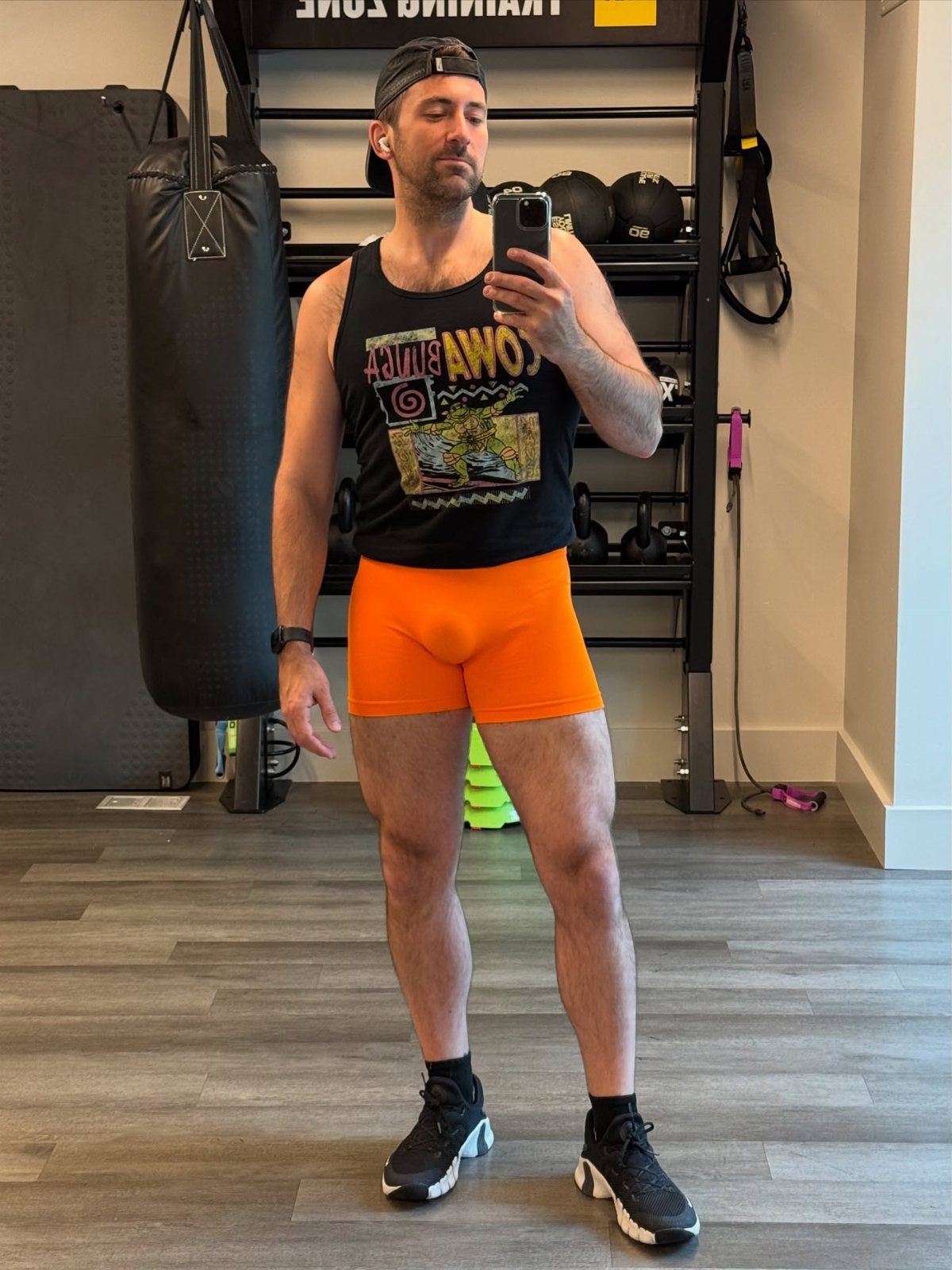 Man in a gym wearing a black tank top and neon orange workout shorts.