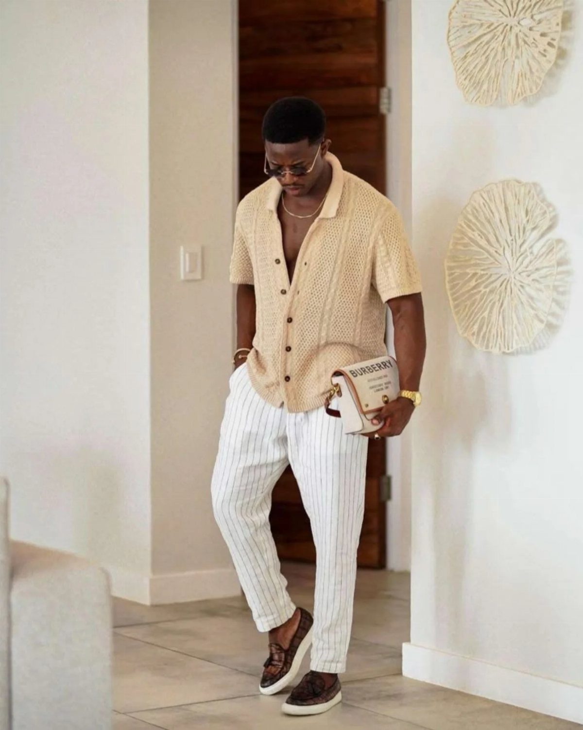 Man in beige knit shirt and white pinstriped trousers holding a white handbag.