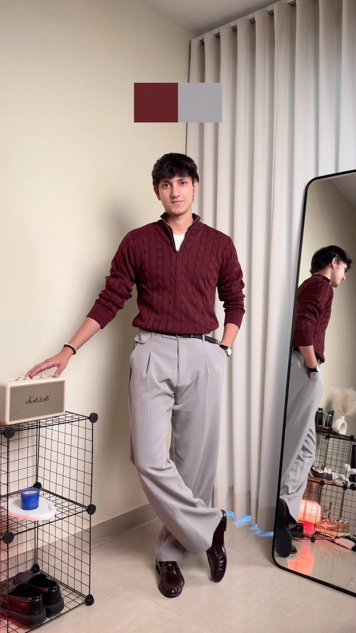 Young man wearing a maroon knit sweater and grey pleated trousers posing indoors.