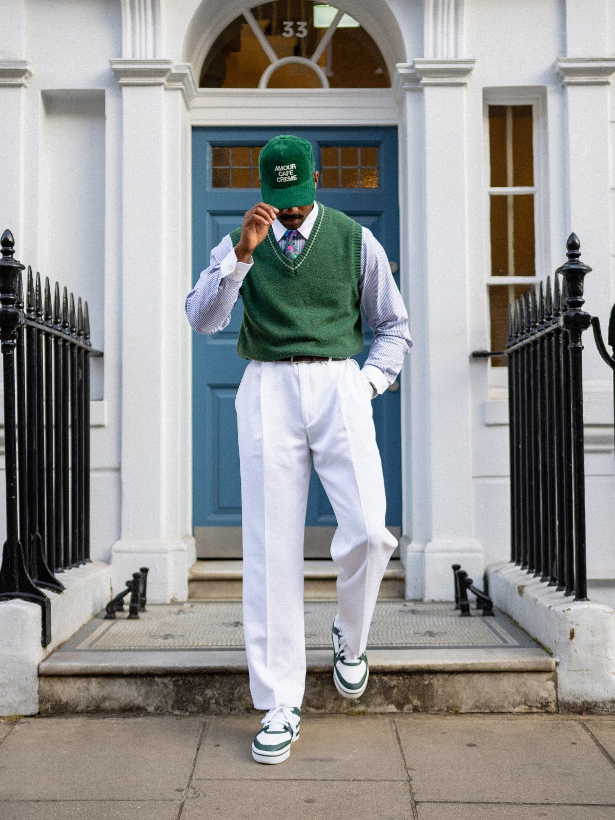 Man wearing a green sweater vest, striped shirt, and white trousers by a door.