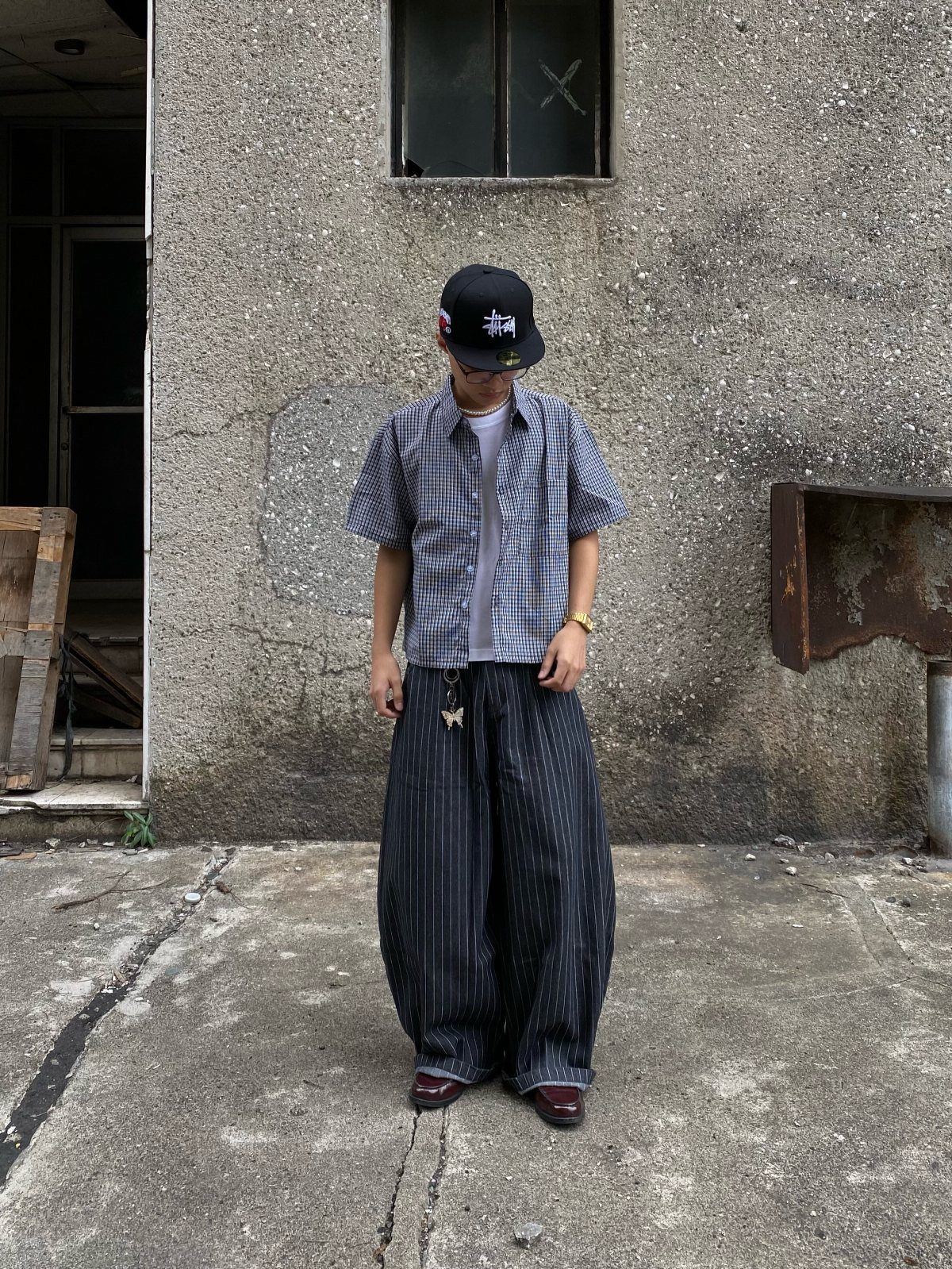 Person in checkered shirt and wide pinstriped trousers standing by a concrete wall