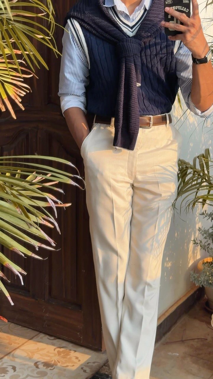 Person wearing a striped shirt, navy sweater over shoulders, and cream pleated trousers.