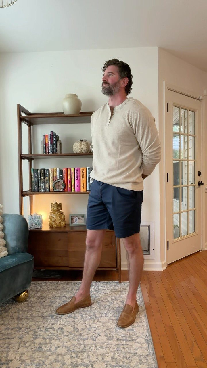 Man wearing a beige henley shirt and navy blue long shorts with brown loafers.