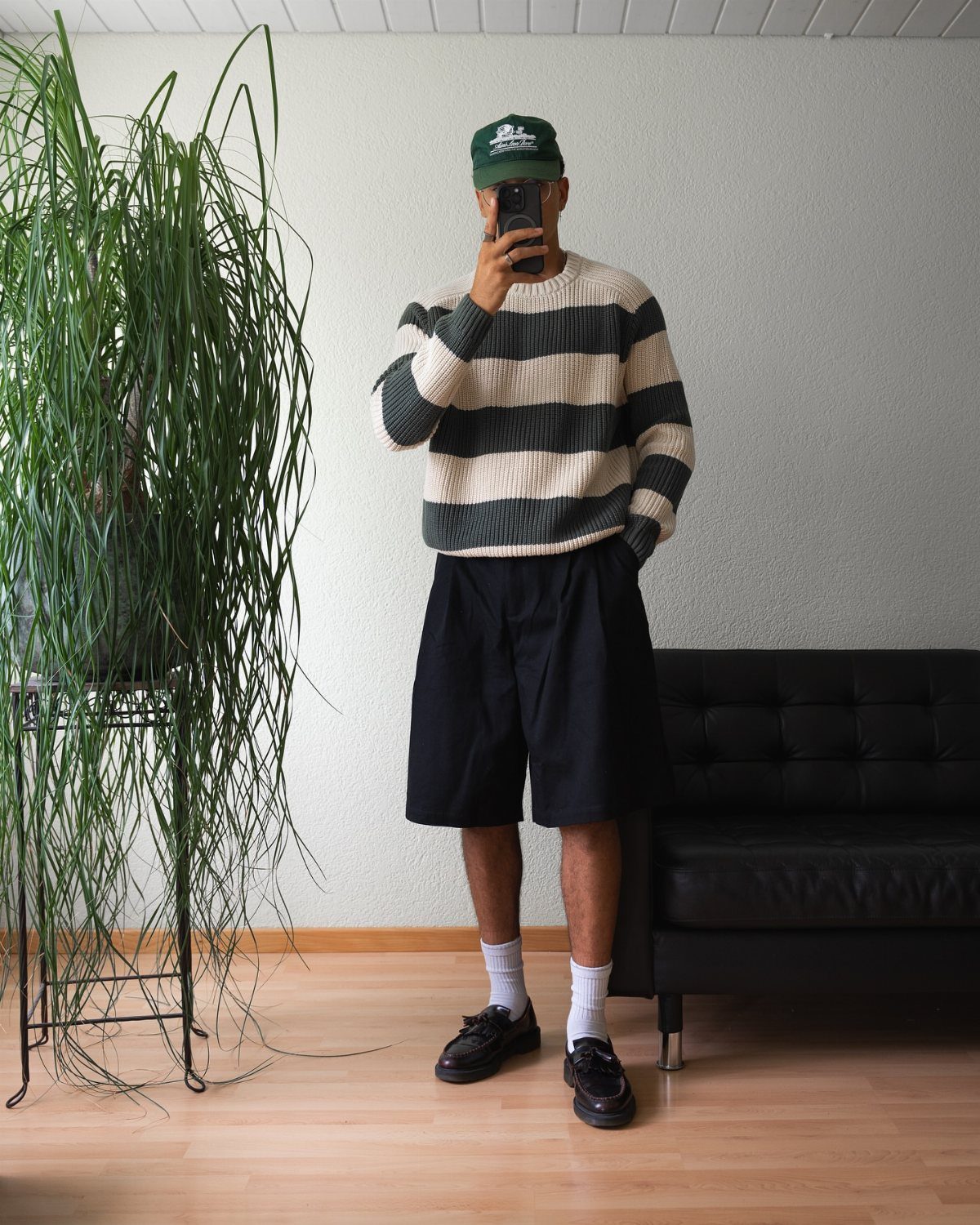 Person wearing a striped sweater, black long shorts, and a green baseball cap.