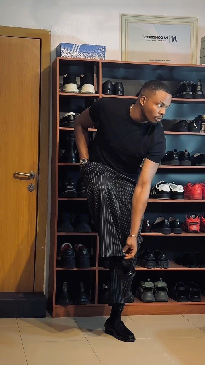 Man in black t-shirt and pinstripe pants posing by a shoe rack.