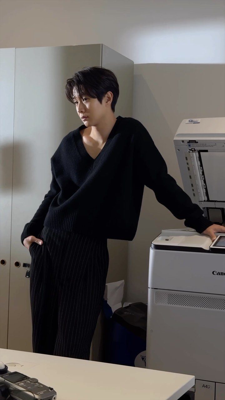 Man in charcoal v-neck sweater and pinstripe pants standing by an office printer.