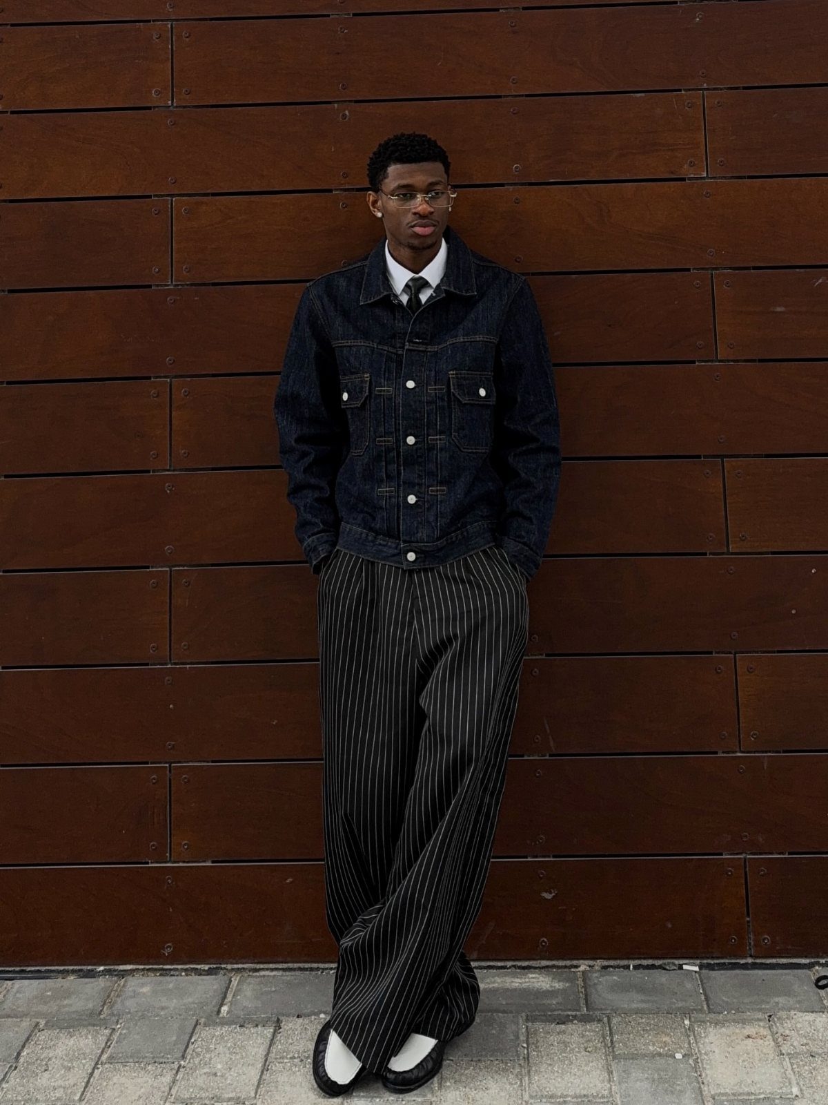 Man wearing a dark denim jacket and wide-leg black pinstripe pants against wood.