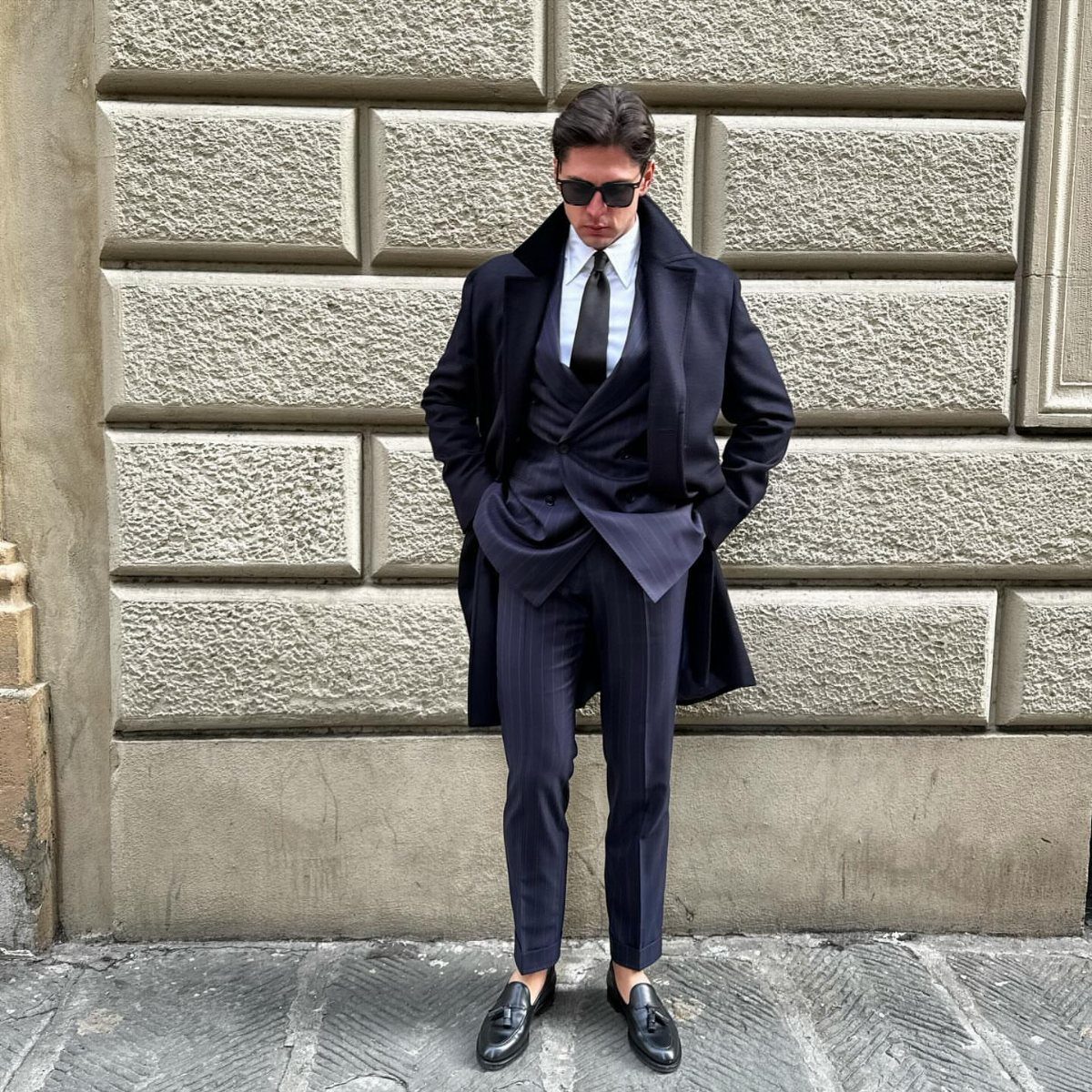 Man in navy overcoat and pinstripe pants standing against a stone wall.