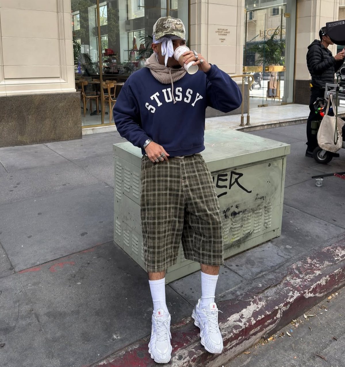 Person wearing navy sweatshirt, olive plaid shorts, and beige scarf on city sidewalk