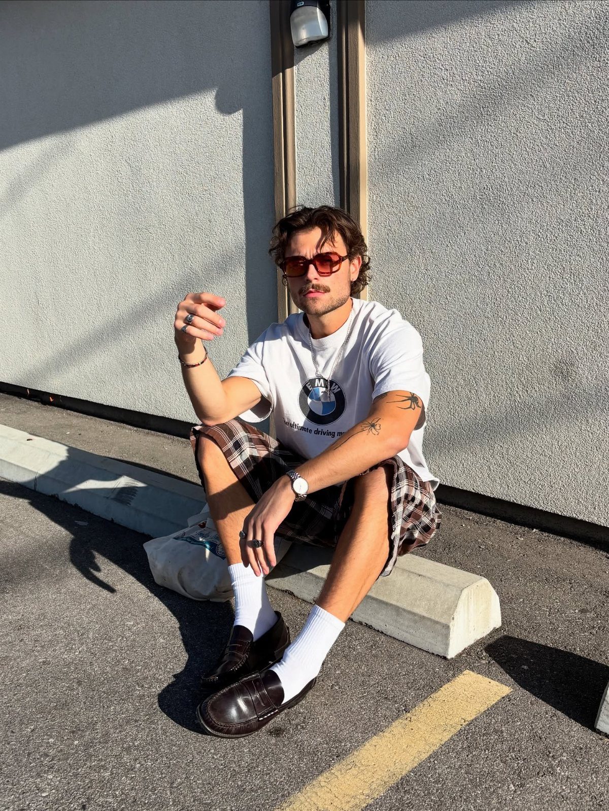 Man wearing white t-shirt, plaid shorts, and red sunglasses sitting on a curb.