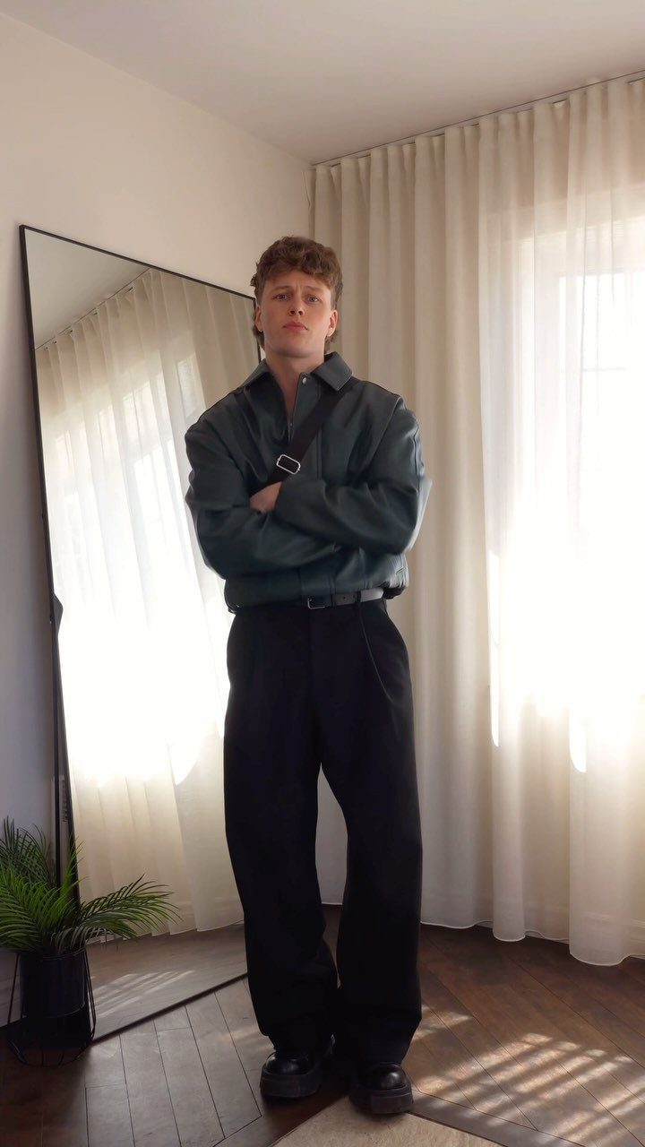 Man wearing a green button-down shirt and wide-leg black pleated trousers in a room.