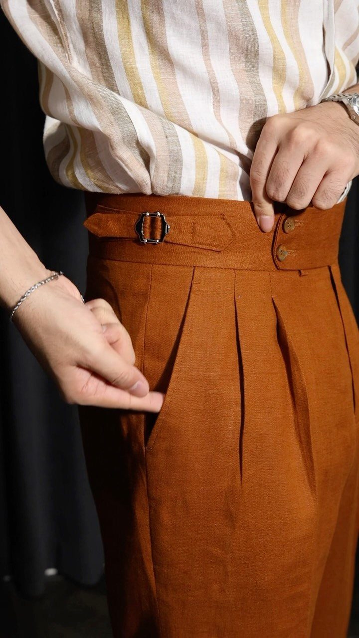 Rust-colored pleated trousers with a side buckle and tucked-in striped shirt.