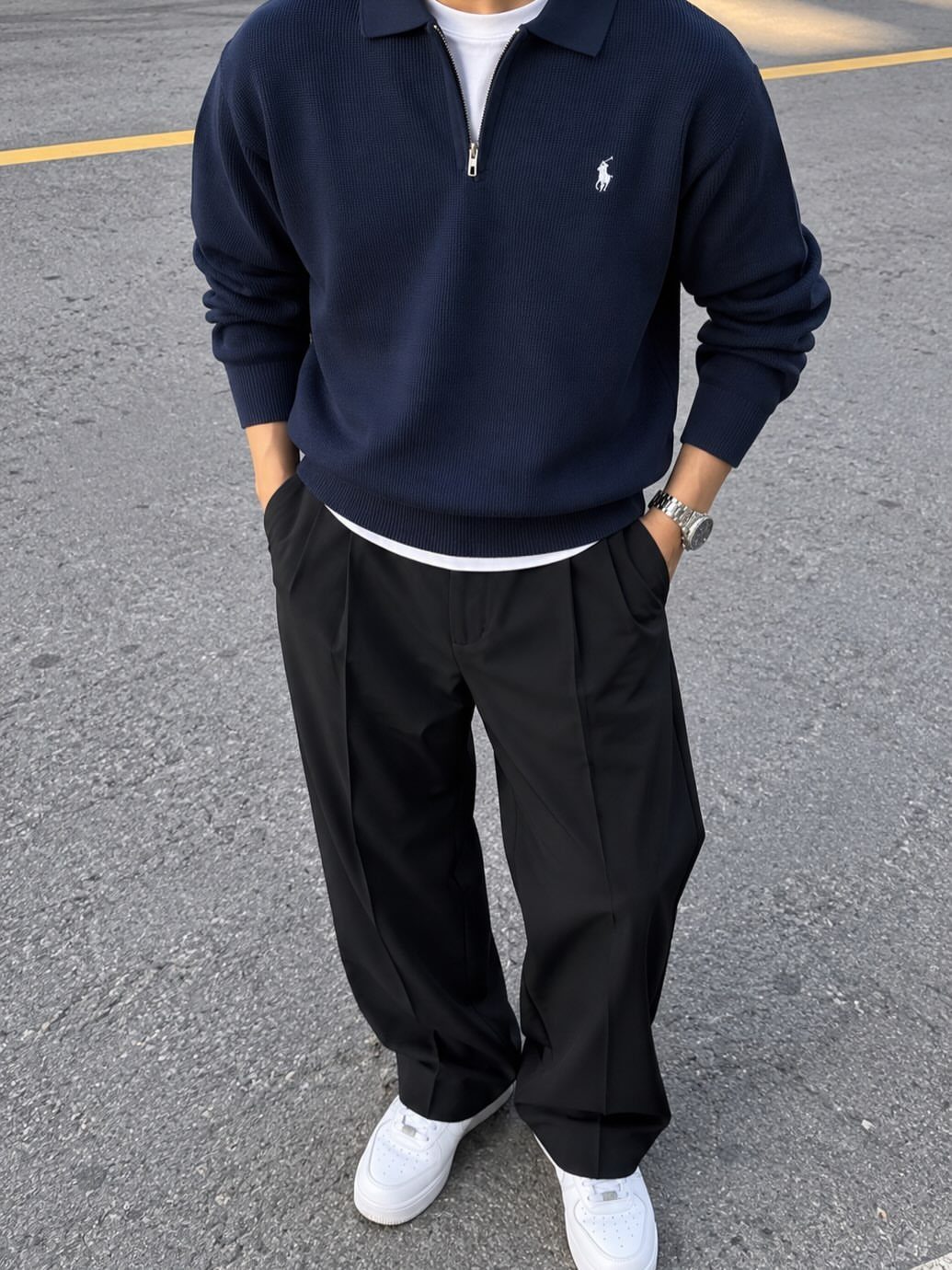 Person wearing navy polo sweater, black pleated trousers, and white sneakers on asphalt.