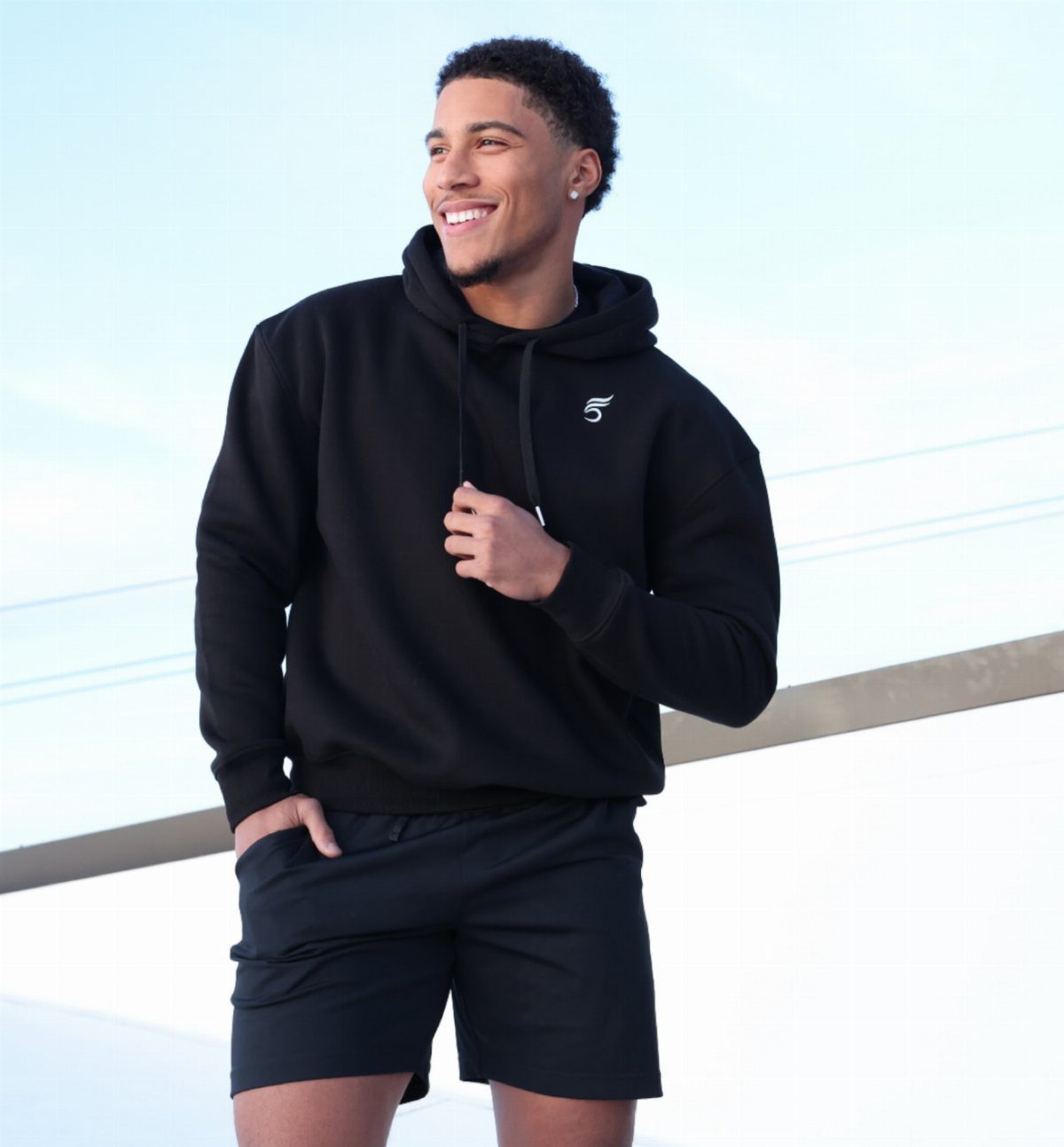 Man wearing a black hoodie and black sports shorts smiling outdoors.