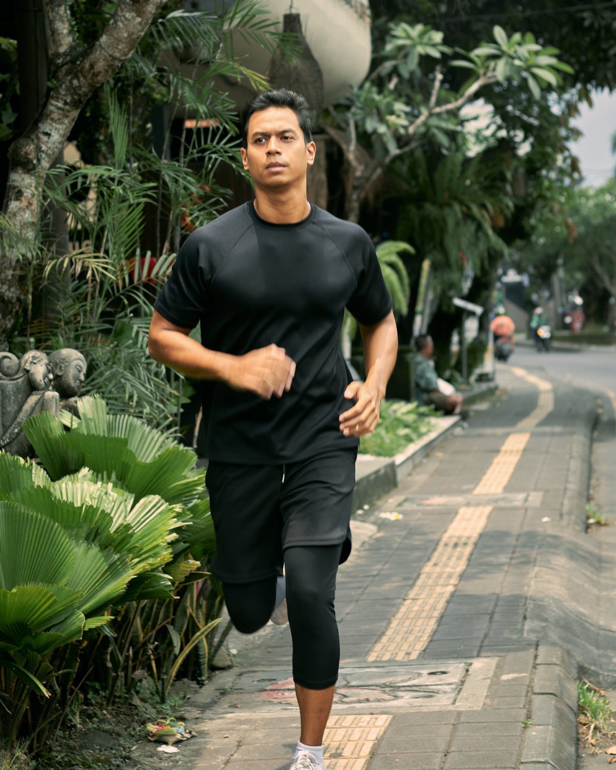 Man running on a sidewalk wearing a black athletic shirt and running shorts.