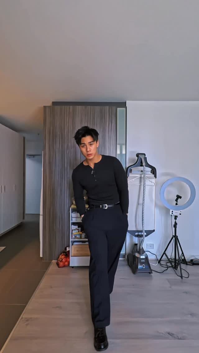 Man wearing a black henley shirt and black trousers posing in a room.