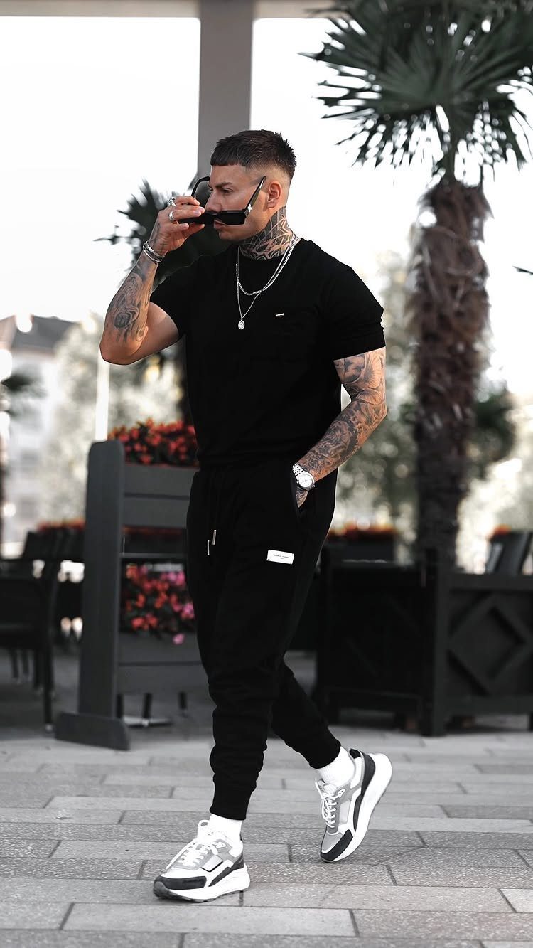 Man in black t-shirt and joggers wearing sneakers and silver chains outdoors.