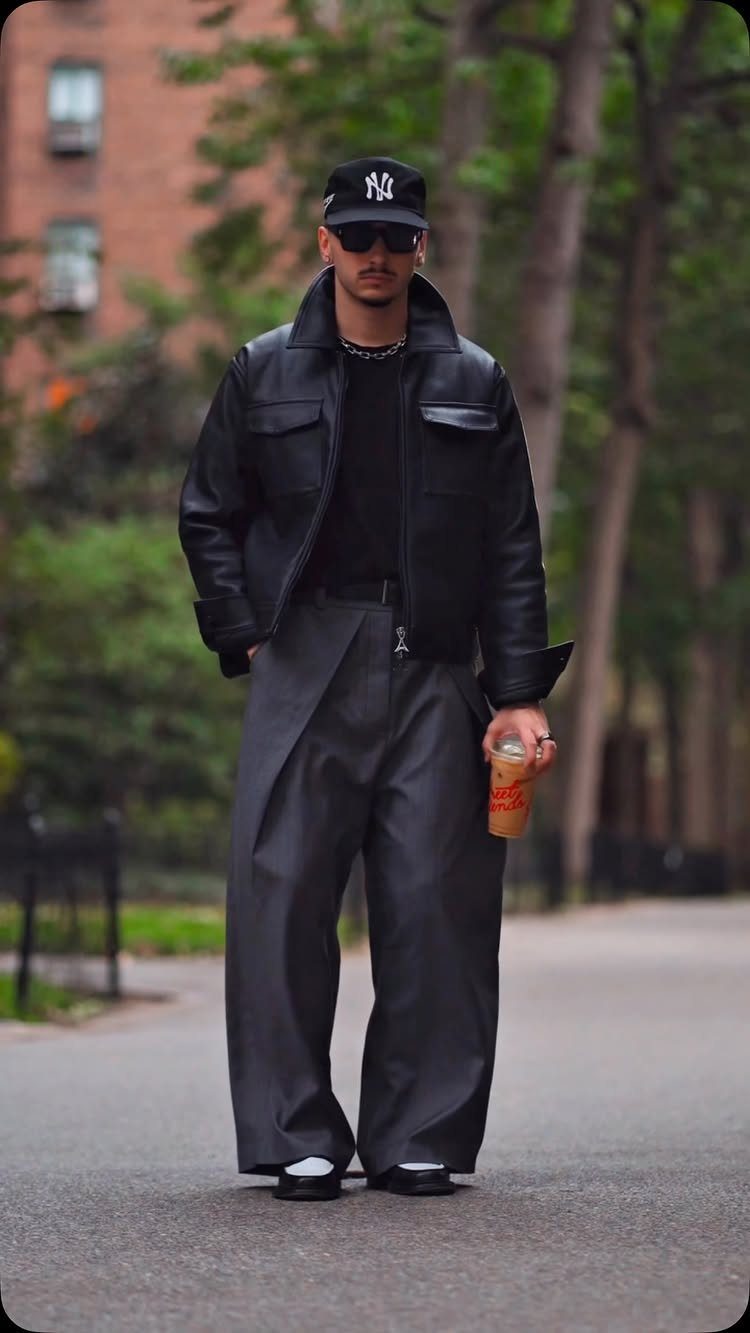 Man wearing black leather jacket, wide grey trousers, and New York Yankees cap.