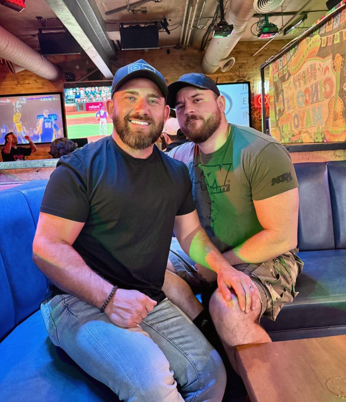 Two men wearing t-shirts and baseball caps sitting in a sports bar booth.