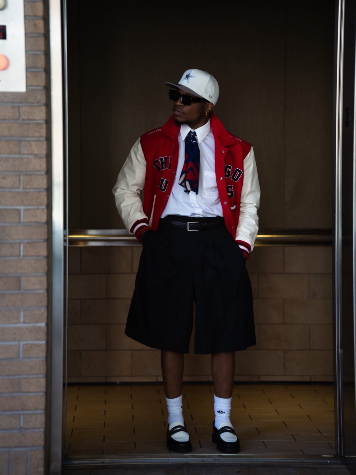 Man in varsity jacket, tie, and baseball cap posing in a doorway.