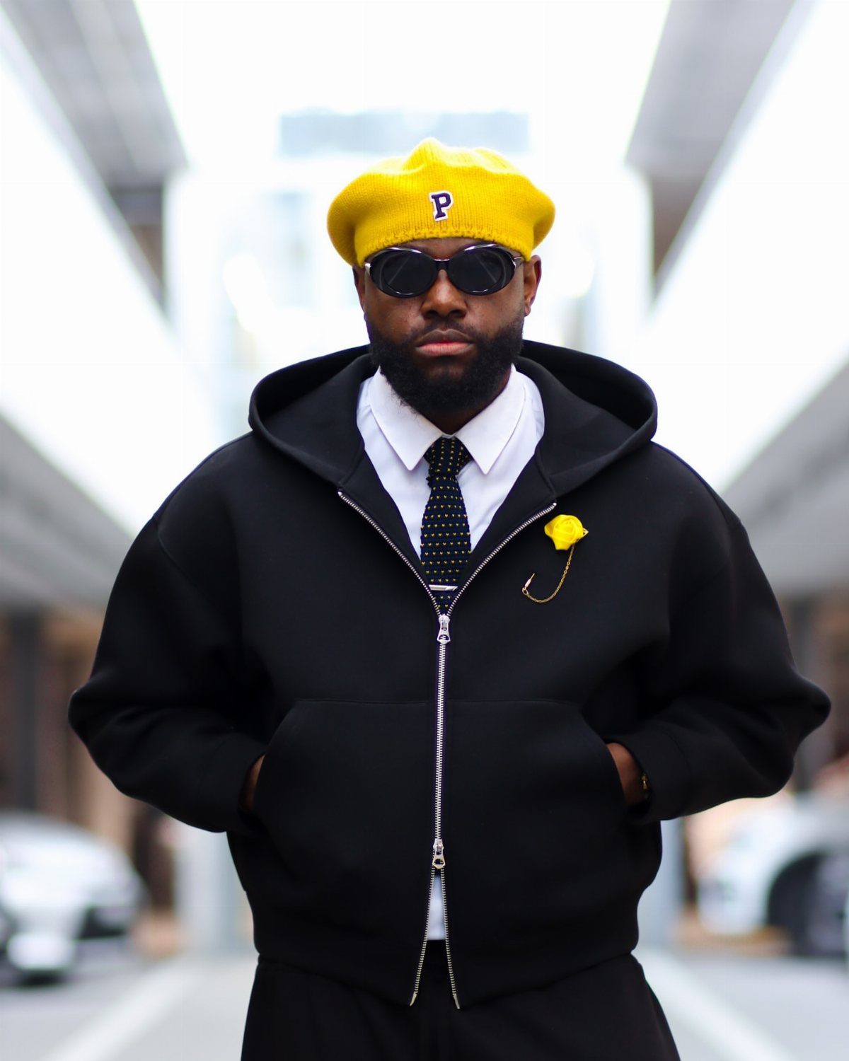 Man in black hoodie, white shirt, black tie, and yellow beret posing outdoors.