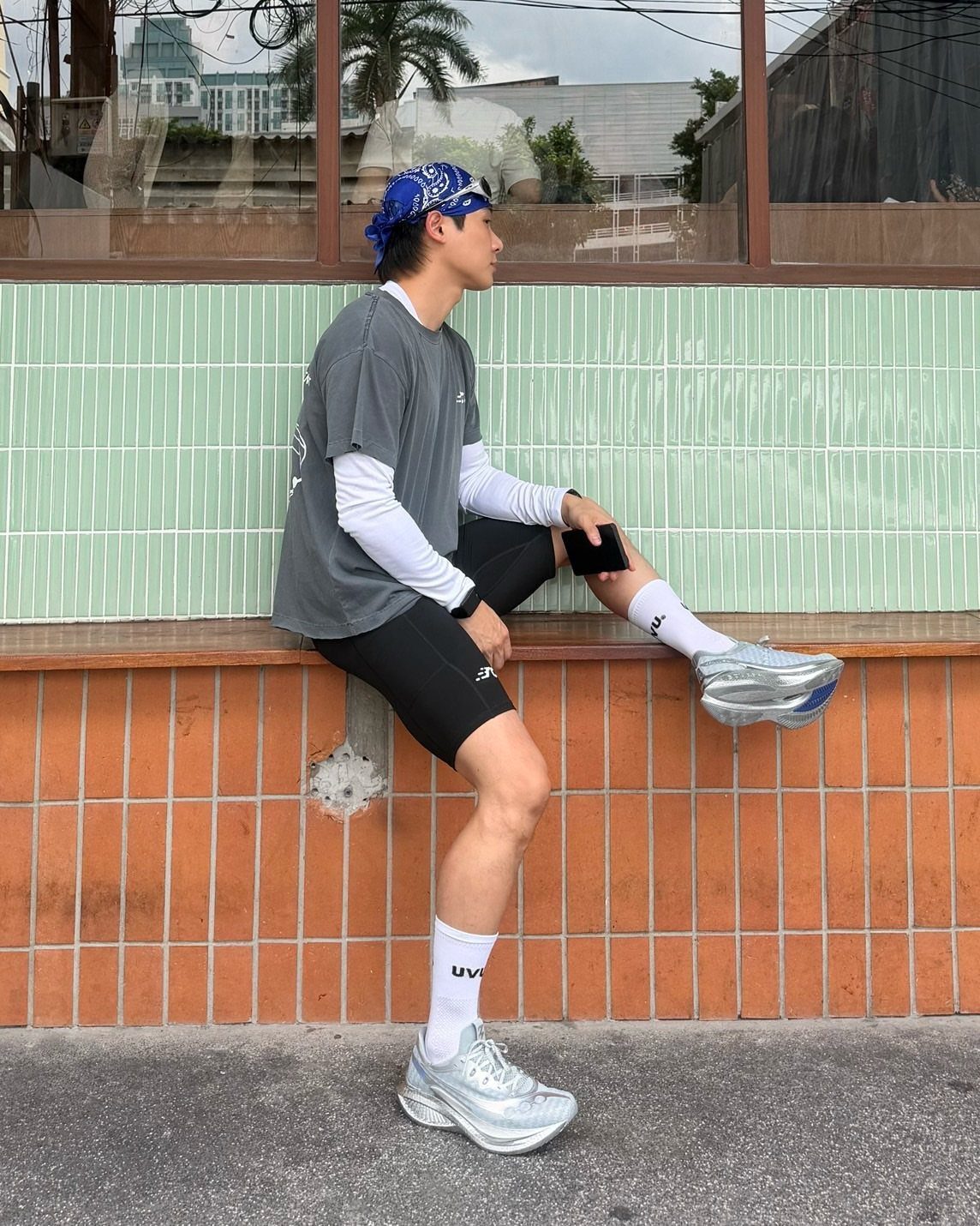 Man in grey t-shirt, black shorts, and silver sneakers posing against tiled wall.