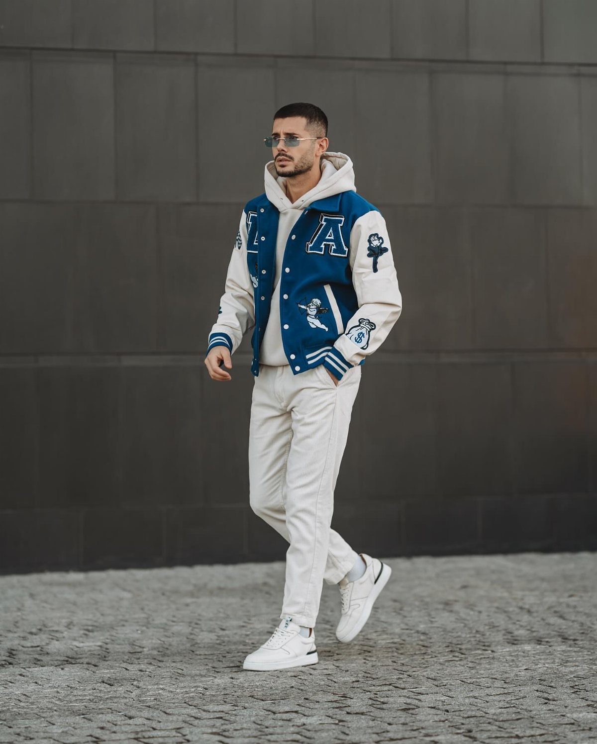 Man wearing a blue and white varsity jacket, white hoodie, and white trousers.