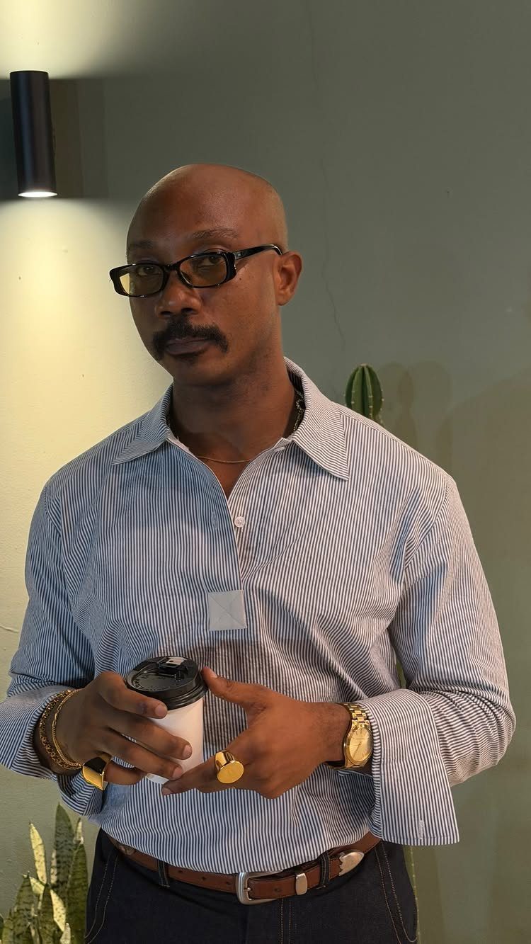 Man wearing a blue striped shirt tucked into dark denim jeans with gold accessories.