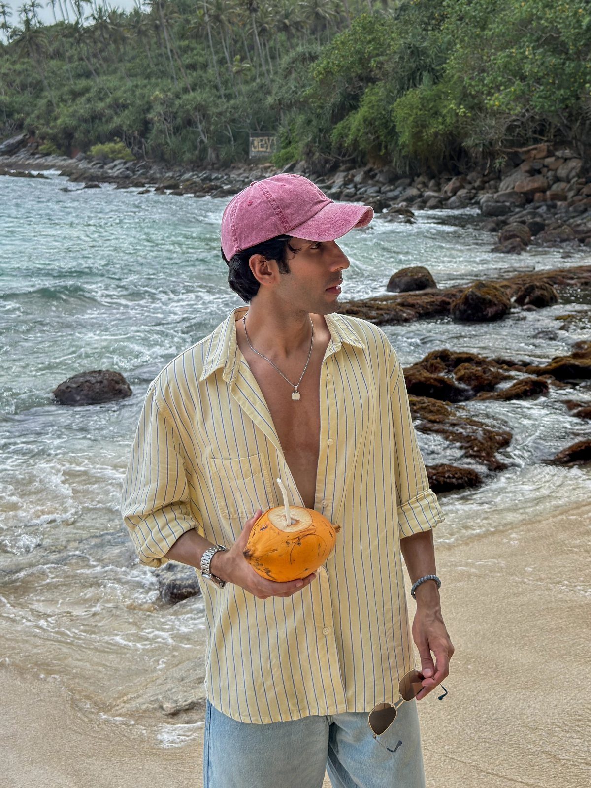 Man in a striped yellow shirt and denim shorts holding a coconut on a beach