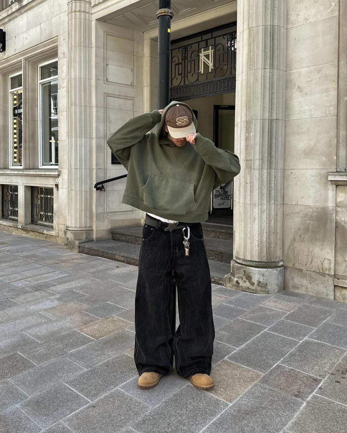Person wearing an olive green hoodie and wide-leg black pants on a city sidewalk.