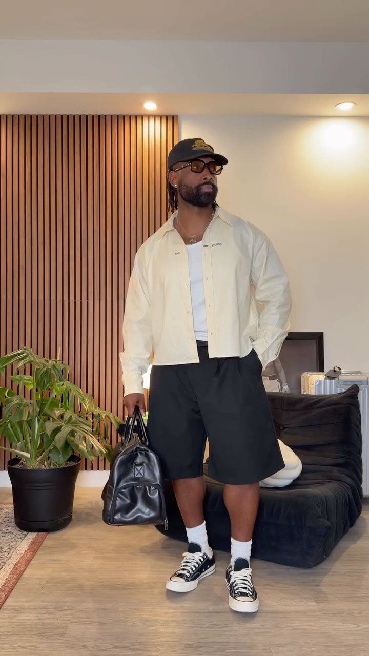 Man wearing cream shirt, loose black shorts, and black sneakers standing indoors.