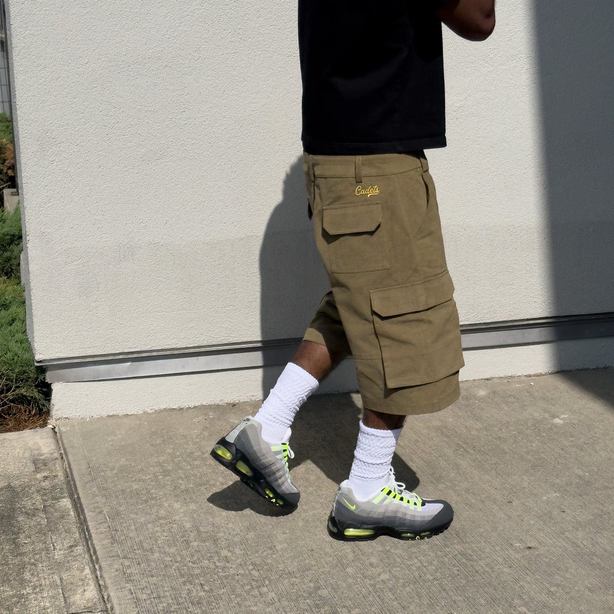 Man wearing khaki cargo shorts, white socks, and grey sneakers against a white wall.